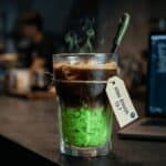 A steaming cocktail with a bright green minty bottom and a tag tied to the rim, placed on a bar beside a laptop showing code in the background.