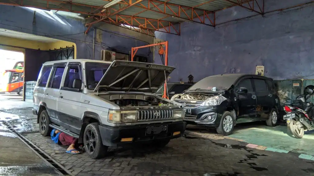 Auto repair shop with a silver suv and a black suv parked inside; hood open on both vehicles, a worker lies under the silver suv under the front wheel area in sandals.