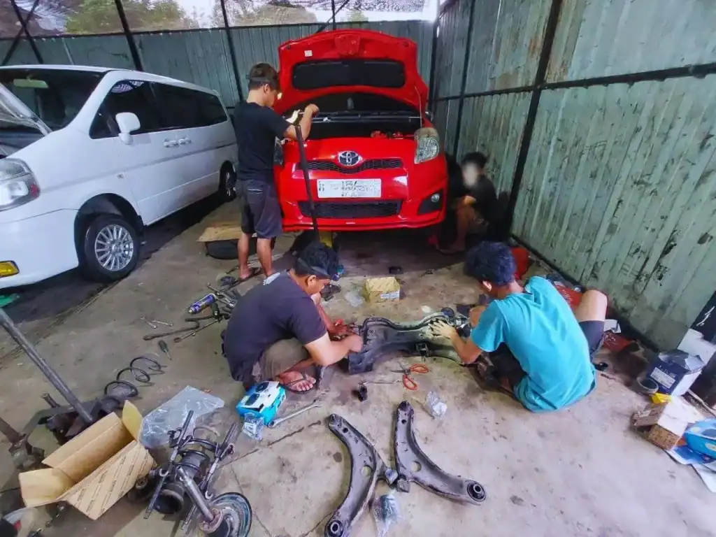 Three mechanics repair a red car with the hood up in a cluttered garage.