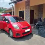 Red compact car with front and passenger doors open parked in a residential driveway; a person’s legs visible by the open door on the left edge. A motorcycles and a gate are nearby. The house has tan walls and brick pillar accents.