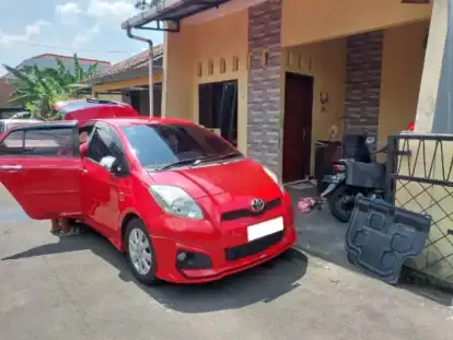 Red compact car with front and passenger doors open parked in a residential driveway; a person’s legs visible by the open door on the left edge. A motorcycles and a gate are nearby. The house has tan walls and brick pillar accents.