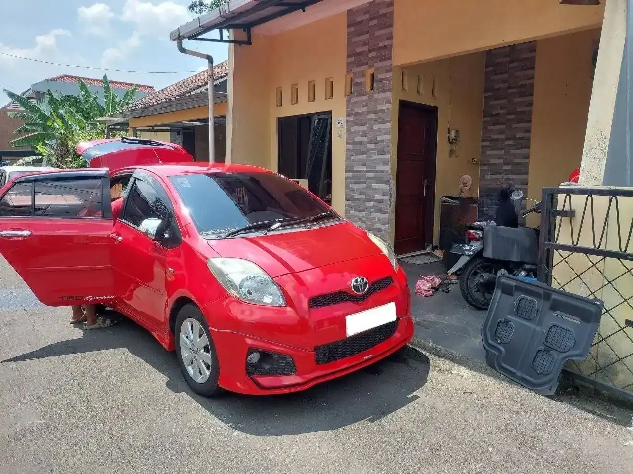 Red compact car with front and passenger doors open parked in a residential driveway; a person’s legs visible by the open door on the left edge. A motorcycles and a gate are nearby. The house has tan walls and brick pillar accents.